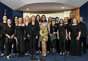 A imagem mostra um grupo de pessoas posando para uma foto em um ambiente formal. A maioria dos participantes são mulheres vestidas de preto, enquanto uma mulher ao centro usa um vestido estampado colorido. O grupo aparenta estar em um evento oficial ou apresentação, possivelmente relacionado à música ou cultura, considerando a presença de partituras e microfones. O cenário tem um fundo azul com detalhes dourados, sugerindo um ambiente institucional ou governamental.