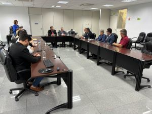 homens e mulheres ocupam uma mesa em U numa sala com piso e teto claros. Eles usam terno