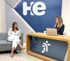 Uma mulher sentada em uma poltrona e um mulher numa bancada. Elas olham para a câmara. Ao fundo, a logomarca do programa hora extra. Embaixo da bancada, a logomarca da Justiça do Trabalho
