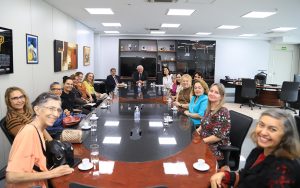 A imagem mostra um grupo de pessoas reunidas ao redor de uma mesa de conferência em uma sala de reunião bem iluminada. A maioria dos participantes são mulheres, todas sorrindo e parecendo engajadas na conversa. Há um homem sentado no centro da mesa, possivelmente conduzindo a reunião. O ambiente tem um aspecto profissional, com prateleiras e quadros na parede, além de materiais como copos de água e papéis sobre a mesa.
