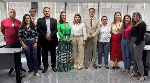 A imagem mostra um grupo de dez pessoas posando para uma foto em um ambiente corporativo. O grupo é composto por homens e mulheres vestidos de maneira formal e semiforma. Alguns usam ternos, enquanto outros estão com roupas sociais mais leves. O ambiente parece ser um escritório ou uma sala de reuniões, com mesas e cadeiras ao fundo. O grupo está organizado em linha, demonstrando um clima profissional e cordial. Todos parecem estar olhando para a câmera e posam de maneira formal.