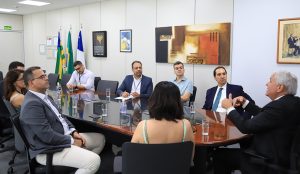 Pessoas sentadas numa mesa oval durante uma reunião. Há quadros fixados na parede