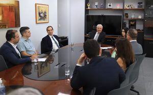 Mesa oval com pessoas sentadas em uma reunião. Há uma estante preta atrás
