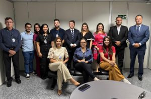 A imagem mostra um grupo de dez pessoas posando para uma foto em um ambiente corporativo. O grupo é compos por homens e mulheres vestidos de maneira formal e semiforma. Alguns usam ternos, enquanto outros estão com roupas sociais mais leves.