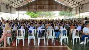 Um ângulo amplo mostra um grande grupo de jovens, provavelmente estudantes, sentados em cadeiras de plástico brancas dentro de um espaço coberto que se assemelha a um ginásio. Eles estão todos sorrindo e segurando pequenos objetos amarelos em suas mãos levantadas. Vários adultos estão sentados entre eles e nas extremidades do grupo, também sorrindo.