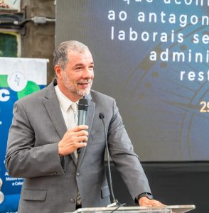 Um homem de pele clara com cabelos grisalhos curtos e barba grisalha rala está falando em um microfone em um pódio transparente. Ele veste um terno cinza, camisa branca e gravata clara com um padrão sutil. Ele olha para a direita e sorri levemente. 