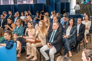 Uma plateia diversificada de pessoas está sentada em cadeiras, olhando em direção à frente, presumivelmente para um apresentador ou evento. As roupas variam de casuais a mais formais, incluindo ternos, blazers e vestidos. 