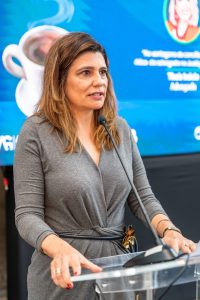 ma mulher de pele clara com cabelos castanhos na altura dos ombros está falando em um microfone atrás de um pódio transparente. Ela veste um vestido cinza de mangas compridas com um cinto escuro na cintura.