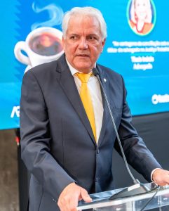 Um homem branco de cabelos brancos e terno azul fala ao microfone em um pódio transparente.