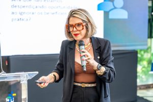 Uma mulher de pele clara com cabelos loiros curtos e óculos grandes de armação marrom está falando em um microfone. Ela veste um blazer preto sobre uma blusa marrom, calças pretas e um cinto branco com uma fivela dourada. 