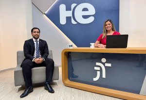 Um homem sentado em uma poltrona e uma mulher numa bancada. Eles olham para a câmara. Ao fundo, a logomarca do programa hora extra. Embaixo da bancada, a logomarca da Justiça do Trabalho
