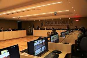 Fotografia ampla de um auditório no TRT-GO, com mesas de videoconferência em primeiro plano e uma plataforma elevada com juízes atrás. A parede exibe "JUSTIÇA DO TRABALHO - Tribunal Regional do Trabalho da 18ª Região (GO)". Bandeiras brasileiras visíveis. 