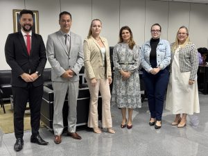 A imagem mostra um grupo de seis pessoas em pé, lado a lado, posando para uma foto em um ambiente interno que parece ser um escritório ou sala de reuniões. Há dois homens e quatro mulheres no grupo.