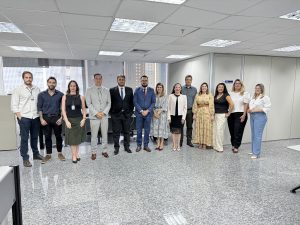A imagem mostra um grupo de doze pessoas em pé, lado a lado, posando para uma foto em um ambiente interno que parece ser um escritório. Há sete homens e cinco mulheres no grupo.
