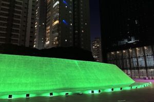 A imagem captura um momento noturno em frente ao prédio do Tribunal Regional do Trabalho da 18ª Região (TRT 18). O prédio está iluminado em verde vibrante, criando um forte contraste com o chão escuro ao seu redor em alusão a campanha do Abril Verde.