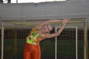 Uma mulher vibrante, vestindo leggings laranjas e uma regata colorida, alonga-se dinamicamente em frente a uma baliza de futebol. Seus braços estão erguidos acima da cabeça, dobrados nos cotovelos, enquanto ela se inclina para a direita com uma expressão alegre no rosto. Ela tem cabelos escuros presos em um rabo de cavalo e parece estar em uma instalação esportiva coberta ou interna bem iluminada, com uma cerca de arame visível ao fundo.