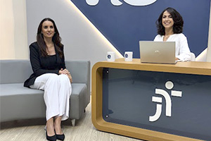 Duas mulheres sorridentes estão em um estúdio de televisão. Uma mulher de cabelos longos e escuros está sentada em um sofá cinza à esquerda, vestindo uma blusa preta, blazer preto e calças brancas. A outra mulher, com cabelos castanhos ondulados na altura dos ombros, está sentada atrás de uma mesa de cor clara à direita, usando uma blusa branca. 