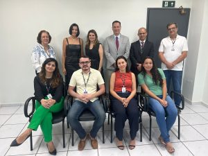 A imagem exibe um grupo de dez pessoas, sendo seis mulheres e quatro homens, posando para uma foto em um escritório. Quatro das pessoas estão sentadas em cadeiras na frente, enquanto as outras seis estão de pé atrás. Ao fundo, há uma parede clara com uma porta escura e um sinal de saída de emergência. O piso é de azulejos claros. As pessoas estão vestidas com uma mistura de trajes sociais e casuais.Fontes