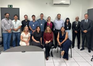 A imagem mostra um grupo de quatorze pessoas, dez homens e quatro mulheres, posando para uma foto em um escritório. Há uma mesa cinza no primeiro plano. Ao fundo, uma parede clara com um ar condicionado, uma porta escura com um sinal de saída e outra porta escura à direita. As pessoas estão vestidas com uma variedade de trajes, desde formais até mais casuais.Fontes