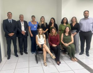 A imagem exibe um grupo de doze pessoas, sendo três homens e nove mulheres, posando para uma foto em um ambiente interno. Duas das mulheres estão sentadas em cadeiras na frente, enquanto as demais pessoas estão de pé atrás. Todos estão vestidos com trajes formais ou semiformais. O fundo é uma parede branca e o piso é de azulejos claros.Fontes