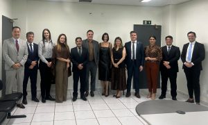 Essa imagem mostra um grupo de doze pessoas, oito homens e quatro mulheres, posando para uma foto em um ambiente interno que parece ser um escritório ou sala de reunião. Todos estão vestidos com trajes sociais. Ao fundo, há uma parede clara com uma porta escura e um sinal de banheiro. Há também parte de uma mesa redonda no canto inferior direito.Fontes