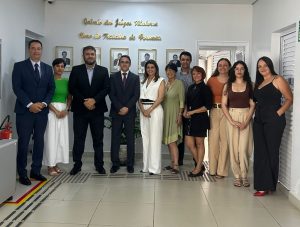 Um grupo de doze pessoas, aparentemente homens e mulheres adultos, posam para uma foto em um corredor bem iluminado. Os homens vestem ternos escuros, camisas e gravatas, enquanto as mulheres usam uma variedade de roupas formais e semi-formais em tons claros e escuros.