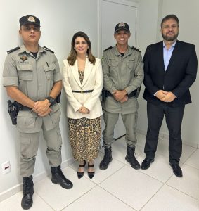 Quatro pessoas posam para uma foto em um ambiente interno. Dois homens à esquerda vestem uniformes militares cinza com quepes e insígnias. Uma mulher no centro veste um vestido estampado de leopardo e um blazer branco. Um homem à direita veste um terno preto sobre uma camisa azul clara.
