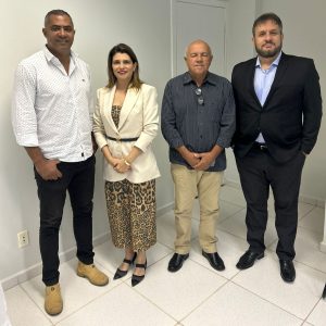 Quatro adultos posam para uma foto em um ambiente interno com paredes claras e piso claro com pequenas manchas escuras. Um homem à esquerda veste uma camisa branca estampada, calças pretas e botas marrons.