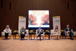 Cinco pessoas estão sentadas em cadeiras no palco de um evento com um telão e banners "5º ENAM" ao fundo.