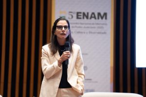 Uma mulher de meia-idade com óculos fala ao microfone em um evento com um banner "5º ENAM" ao fundo.