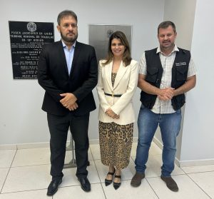 Três pessoas posam em frente a placas com texto em português. Dois homens de terno e uma mulher de vestido e blazer sorriem para a câmera.