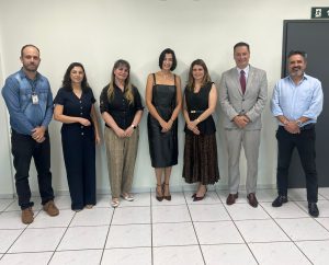 A imagem apresenta um grupo de sete pessoas, sendo quatro mulheres e três homens, posando para uma foto em um ambiente interno. Ao fundo, uma parede clara e, no canto superior direito, um sinal de saída de emergência. O piso é de azulejos claros. As pessoas estão em diversas vestimentas, desde social completo até casual com camisa jeans.Fontes