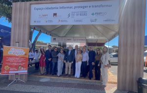 Um grupo de onze pessoas está posando para uma foto em frente a uma estrutura de madeira que parece ser uma espécie de estande ou palco ao ar livre. A estrutura tem uma faixa no topo com a inscrição "Espaço cidadania: informar, proteger e transformar", juntamente com logotipos de várias organizações, incluindo "Justiça do Trabalho TRT-18ª Região", "MPT", "FSSTGO" e outros.