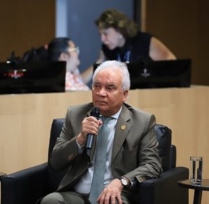 Homem de terno marrom fala ao microfone, sentado em poltrona preta. Mesas laterais com flores e água. Ao fundo, pessoas desfocadas.