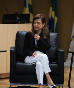 Mulher de blazer preto e calça branca fala ao microfone, sentada em poltrona preta entre mesas com flores. Bandeiras do Brasil e Goiás ao fundo.