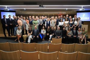 Grande grupo de pessoas posa para foto em auditório com telas "JUSTIÇA DO TRABALHO" ao fundo e cadeiras de madeira vazias à frente.