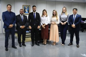 Um grupo de sete pessoas, quatro homens e três mulheres, posa para uma foto em um escritório. Todos estão vestidos com trajes sociais. Ao fundo, há uma parede clara com dois quadros pendurados e parte de uma mesa de escritório.Fontes