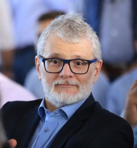 Um homem de com cabelos e barba grisalhos, vestindo óculos azuis, camisa azul e blazer preto, olha para a câmera em um ambiente desfocado com outras pessoas.