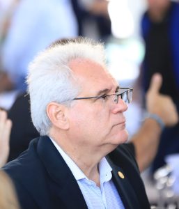 Um homem de perfil, com cabelos brancos e óculos, veste um blazer escuro sobre uma camisa azul clara com listras.