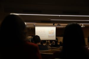 A imagem mostra uma plateia assistindo a uma apresentação em um auditório. No fundo, é visível uma tela de projeção com o título “Igualdade Racial na Tela” e a divulgação do filme “Doutor Gama”.