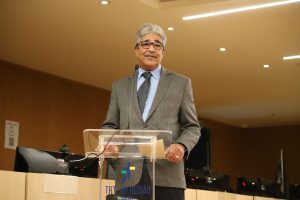 Na imagem, um homem maduro, de cabelos grisalhos e óculos, está em pé atrás de um púlpito transparente, aparentemente discursando.