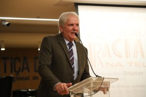 A imagem mostra um homem de cabelos brancos, vestindo terno escuro com camisa clara e gravata listrada, falando ao microfone posicionado em um púlpito transparente.