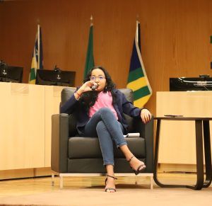 A imagem mostra uma mulher sentada em uma poltrona preta sobre um palco, em um ambiente institucional.