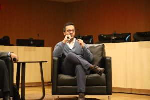 A imagem mostra um homem sentado em uma poltrona preta, participando de uma conversa ou painel em um ambiente institucional. 