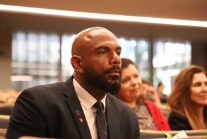 A imagem mostra um homem sentado em um auditório, com expressão atenta e séria, aparentemente concentrado no que está sendo apresentado. 