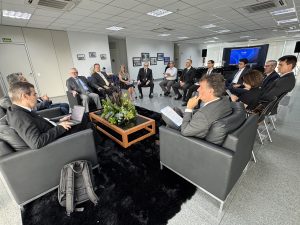Um grupo de pessoas vestidas formalmente participa de uma reunião em uma sala moderna com cadeiras de couro, uma mesa de centro com um arranjo floral.