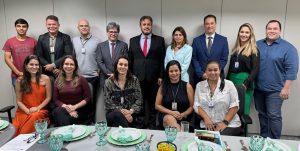 Foto mostra a equipe da 3ª VT de Goiânia com a corregedora do TRT-GO. Algumas pessoas estão sentadas, diante de uma mesa posta com pratos e talheres, e um outro grupo de pessoas está em pé, atrás. Todos posam para a foto.