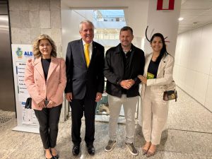Uma foto de corpo inteiro de quatro pessoas, duas mulheres e dois homens, em pé em um corredor. A mulher à esquerda veste um blazer rosa claro e calças escuras. Ao lado dela, um homem de terno escuro e gravata amarela. O outro homem veste um casaco escuro, camisa polo cinza e calças claras. A mulher à direita veste um blazer claro, uma blusa escura e calças claras. Atrás deles, há um sinal com a palavra "BLOCO" e uma lista de salas.