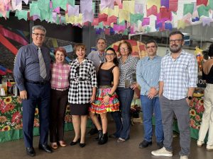 A imagem mostra um grupo de nove pessoas, aparentemente em um evento, com decorações festivas ao fundo.