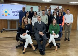 A imagem mostra um grupo de cerca de 20 pessoas posando para uma foto em um ambiente corporativo, provavelmente uma sala de reuniões ou treinamento. Três pessoas estão sentadas à frente, sendo duas mulheres e um homem (ao centro), e os demais estão em pé ao fundo, formando duas fileiras. Todos estão vestidos de forma formal ou social. 